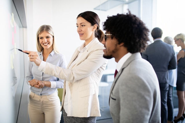 Three people in business attire are discussing at a whiteboard in an office, with others in the background. They appear engaged and focused.