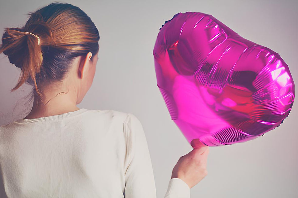 A woman with her hair tied back holds a shiny pink heart-shaped balloon, facing away from the camera against a plain background.