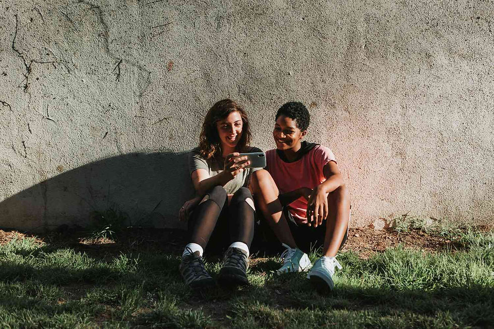 Two people sit against a sunlit wall, smiling at a phone. The scene is grassy, bright, with relaxed, joyful vibes.