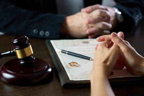 Hands folded over a document with rings, a pen, and a gavel nearby. Two people are seated in a solemn setting.