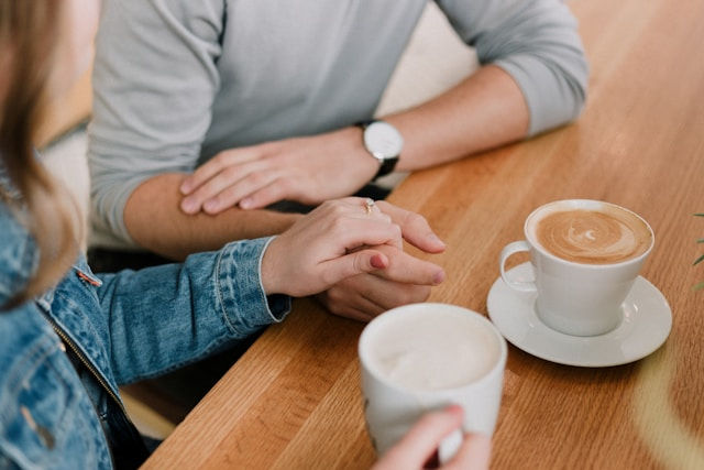 Two people hold hands across a wooden table with two cups of coffee. One wears a denim jacket, creating a warm, cozy atmosphere.