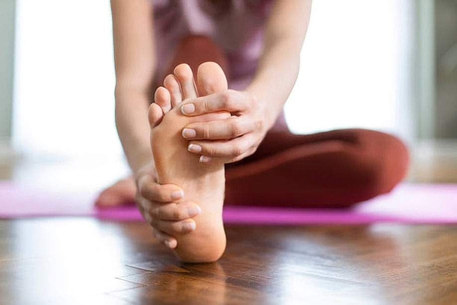 Person in workout clothes sits on a pink yoga mat stretching their foot with both hands. The floor is wooden, and the setting is bright.