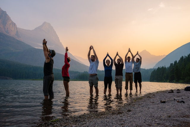 A group of people standing in shallow water near a lakeshore at sunset, performing yoga or stretching poses with a scenic mountain backdrop.
