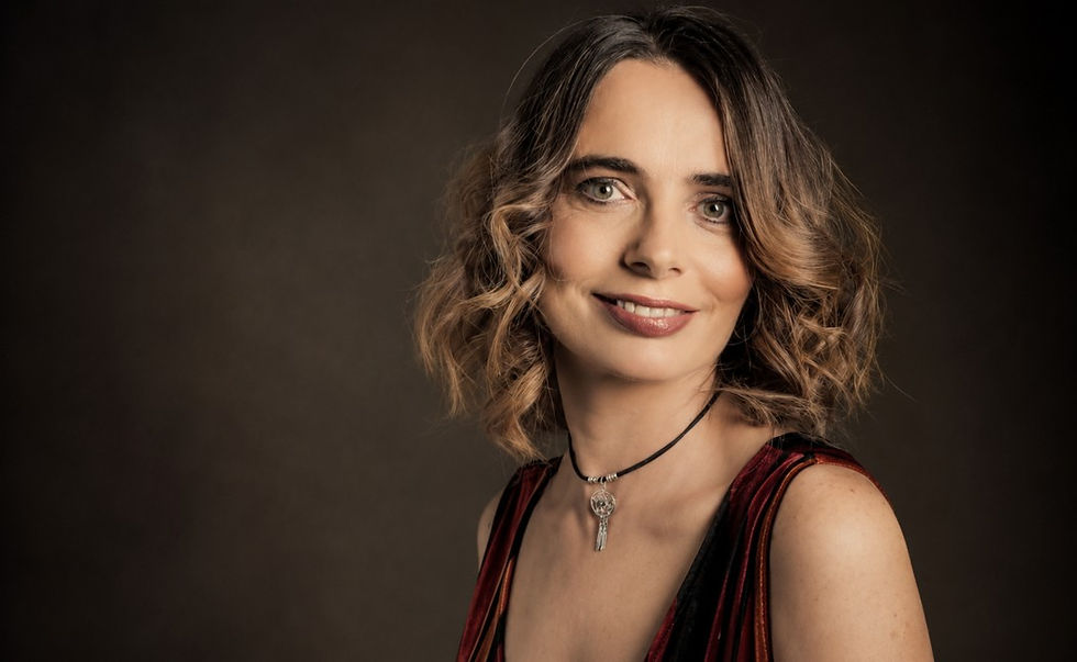 Smiling woman with wavy hair and pendant necklace poses against a dark studio background.