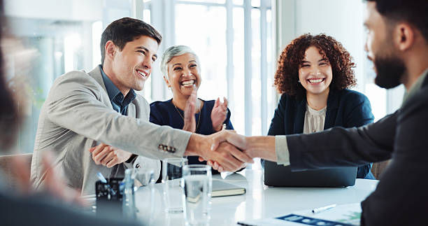 Business meeting in a bright office, two men shaking hands. Three colleagues smiling, one clapping. Mood is positive and collaborative.