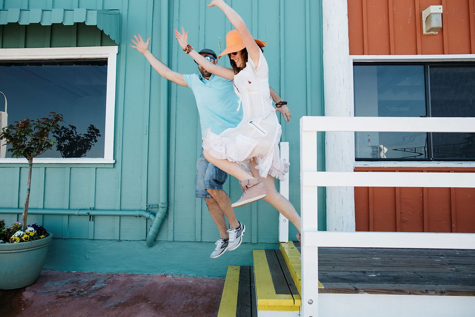 A man in blue and a woman in white with an orange hat joyfully jump off teal steps in front of a colorful building.