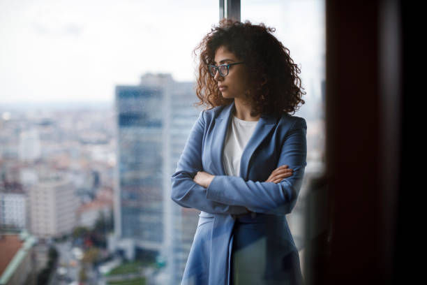 Woman in a blue blazer with curly hair gazes out a window in a high-rise, arms crossed. Urban cityscape visible in the background.