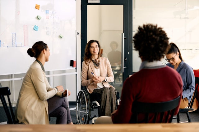 Four people in an office setting having a discussion. One person is in a wheelchair, holding a pen. A whiteboard with charts is visible.