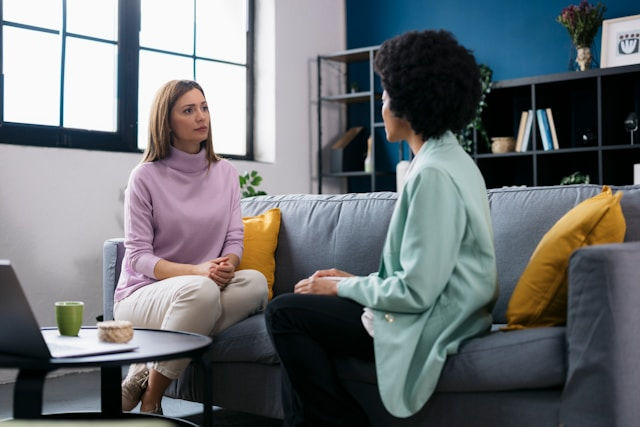 A therapist and a client sit on a couch engaged in a focused conversation during a counseling session in a bright, modern office.