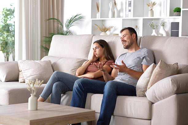 Man and woman sitting on beige sofa in living room, man talking animatedly, woman appears bored or annoyed. Neutral-toned decor.