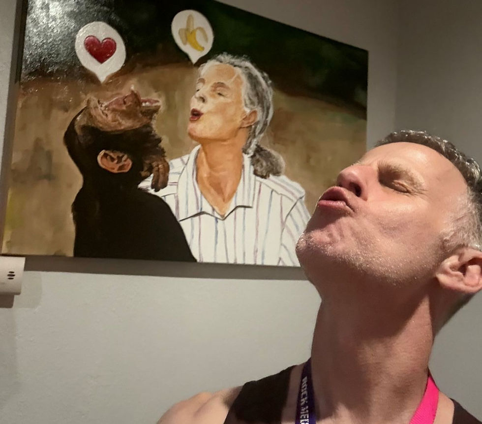 Man posing in front of a painting that shows a chimpanzee and an older woman making kissing faces, with heart and banana thought bubbles above them.