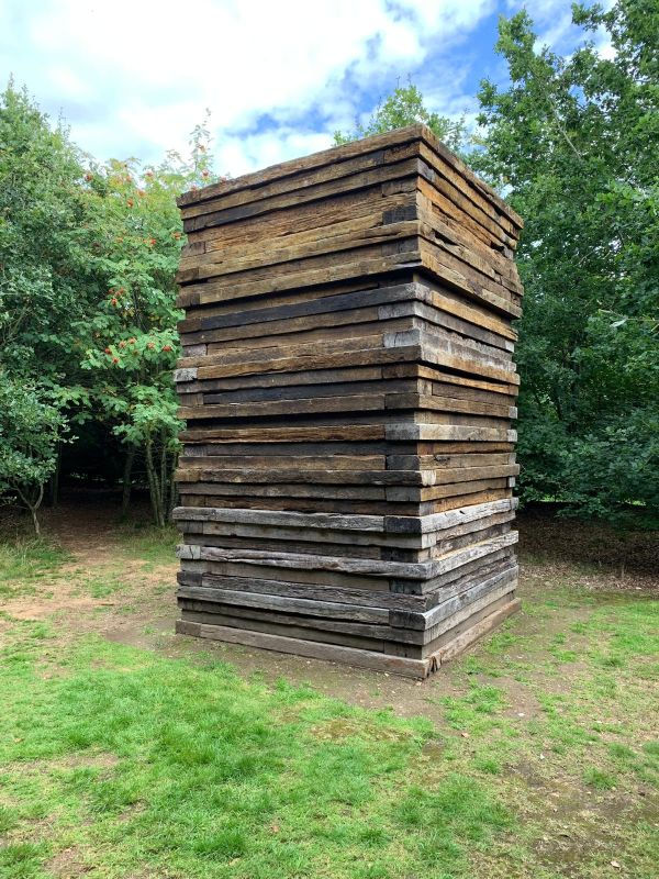 Tall stack of weathered wooden planks in a grassy clearing, surrounded by trees under a cloudy blue sky.