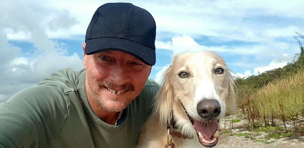 Man in a cap smiles beside a happy dog on a grassy hillside. Bright blue sky and clouds in the background create a cheerful mood.