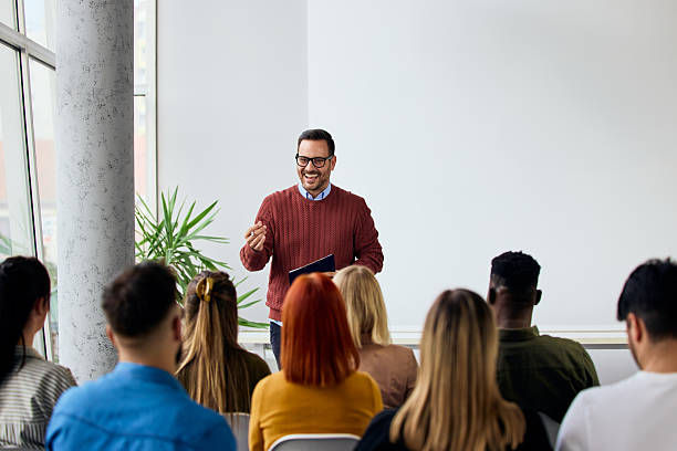 Smiling man in red sweater presents to a seated audience in a bright meeting room with white walls and a plant.