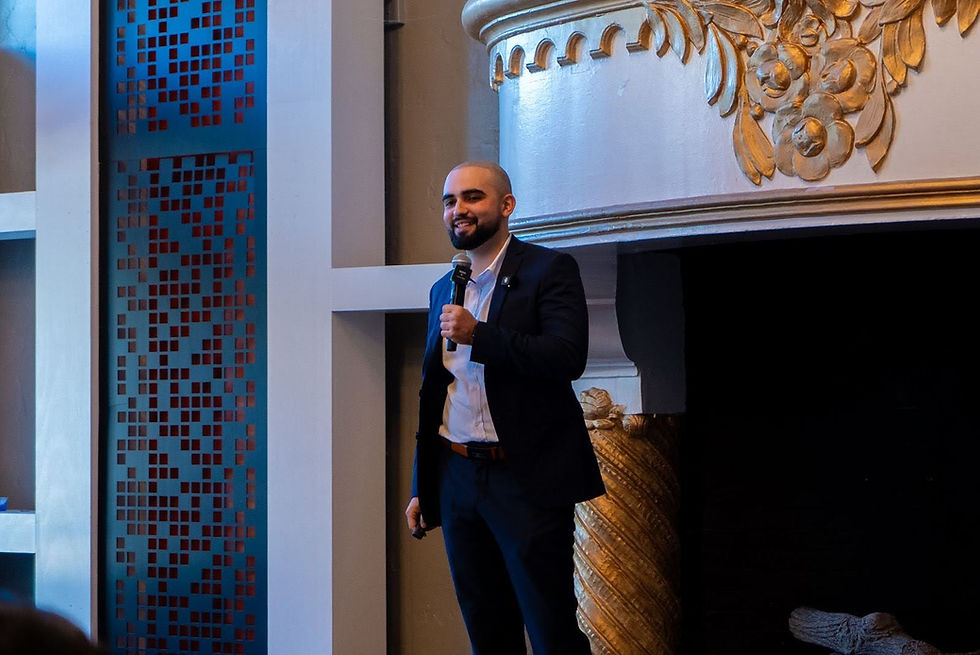 Man in a suit holding a microphone, smiling, standing near an ornate fireplace. Blue patterned panel in the background; warm, elegant setting.