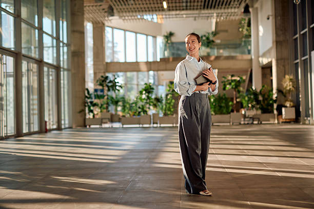 Woman holding a laptop smiles confidently in a sunlit, spacious office with large windows and plants. She wears a light shirt and dark pants.