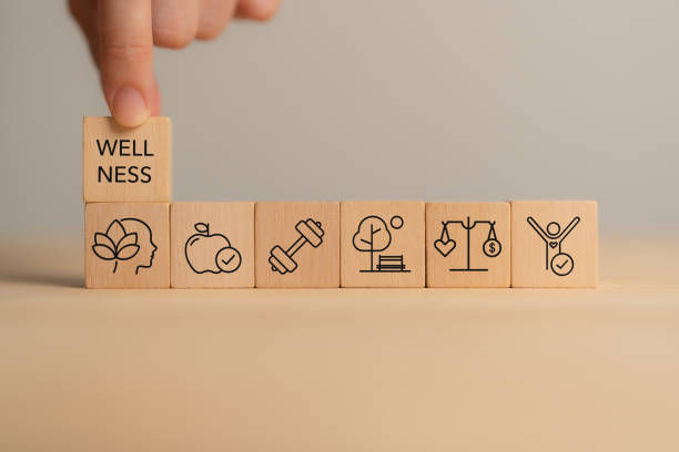 Hand arranges wooden blocks displaying icons for wellness aspects, including exercise and nature. One block reads "WELLNESS." Neutral background.