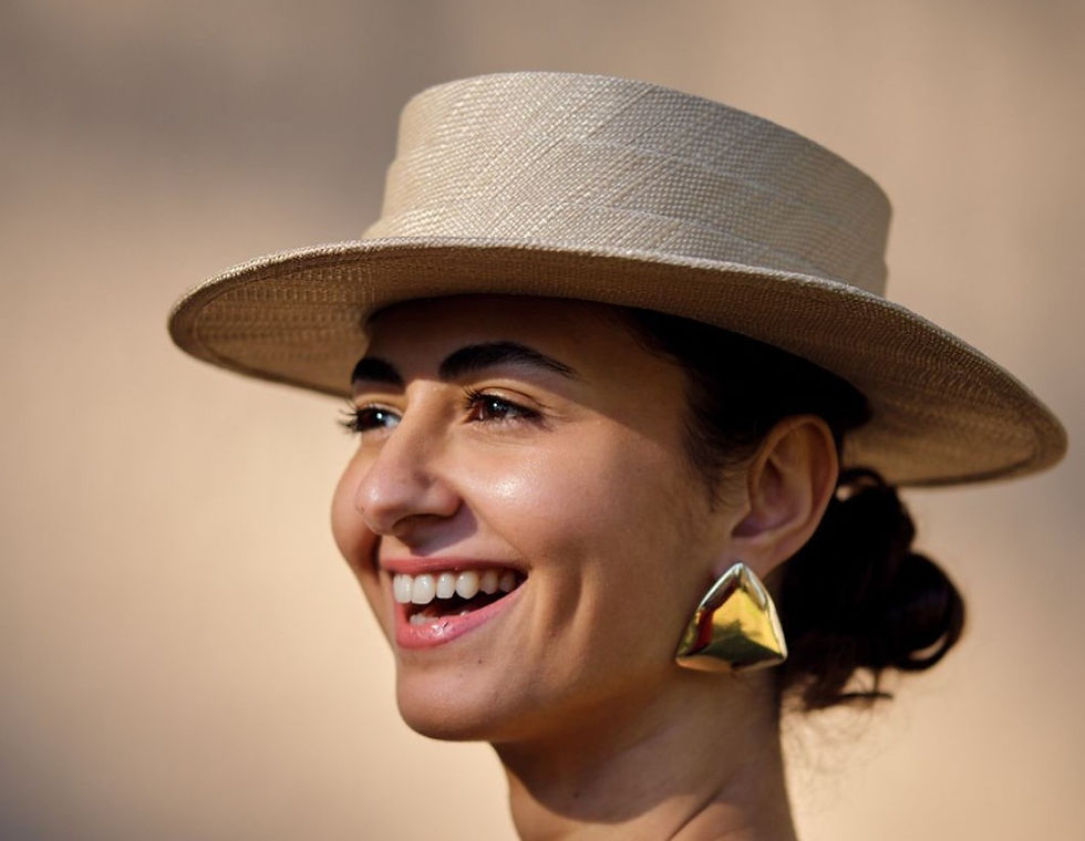Smiling woman wearing a beige straw hat and gold geometric earrings against a soft, neutral background. Mood is joyful and relaxed.