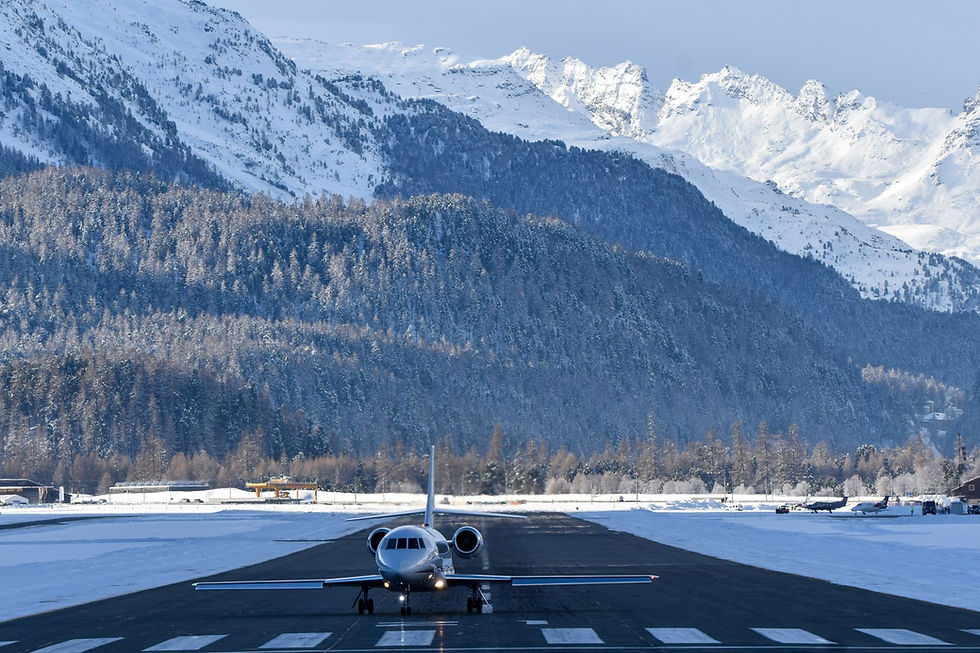 Private jet on a snowy runway with headlights on, framed by snow-covered mountains and forest under a clear blue sky.