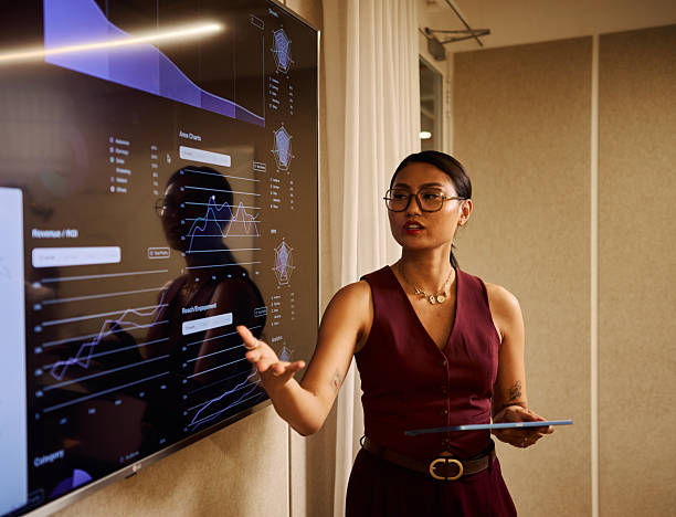 Woman in burgundy dress presents data charts on a wall screen in a conference room, holding a tablet and gesturing confidently