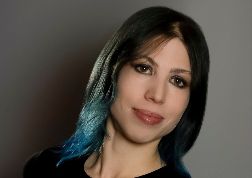 Smiling person with black and blue hair against a gray background. The mood is friendly and the lighting is soft.