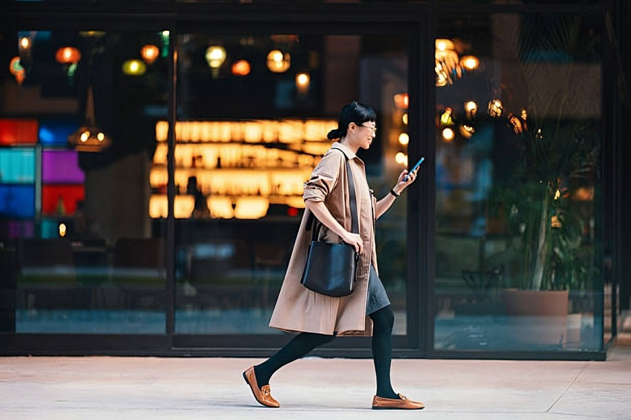 Woman in beige coat walks while looking at phone. She's in front of a bright, colorful cafe window, with a casual and focused mood.