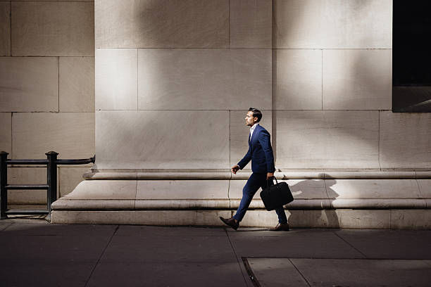 Man in a blue suit walks with a briefcase past a sunlit stone wall, casting a long shadow on the sidewalk.