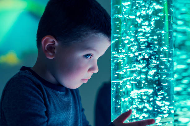Young boy gazes intently at a bubbling blue-lit water tube in a dimly lit room, creating a calm and curious mood.