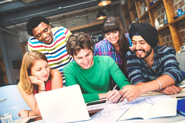 Five people smile and collaborate on paperwork around a laptop in a cozy, book-filled room. They're focused and engaged in discussion.