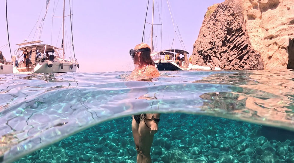 Woman snorkeling in clear turquoise water near sailboats and a rocky cliff under a sunny pink sky.