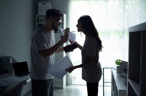 Couple arguing in a bright apartment, man holding cash and a wallet, woman holding papers; tense, backlit scene.