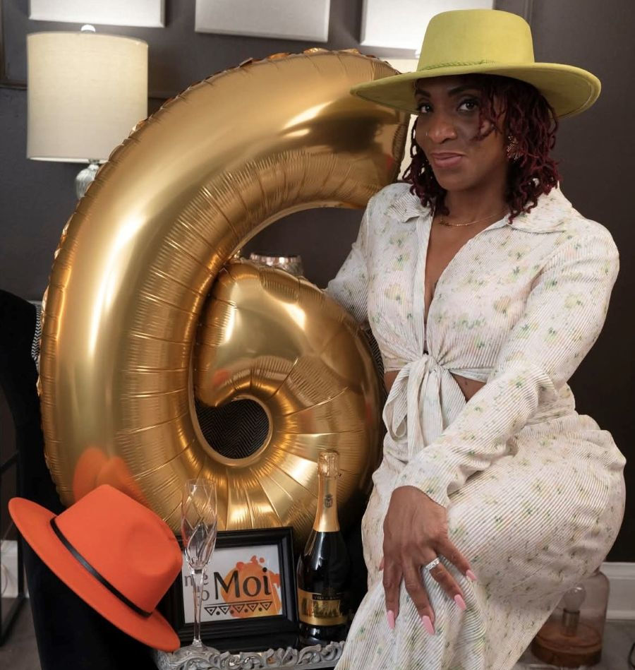 A woman in a light floral dress and green hat poses beside a large gold number 6 balloon, with champagne, glasses, and a framed “Moi” sign on the table.