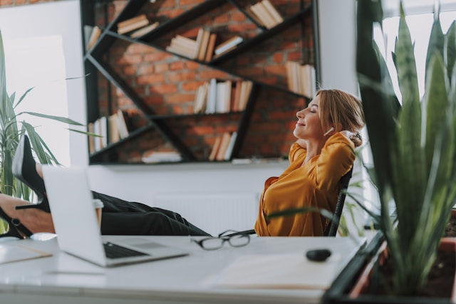 Woman in a mustard sweater reclines in a modern office, stretching beside a laptop, plants, and bookshelves, looking relaxed.