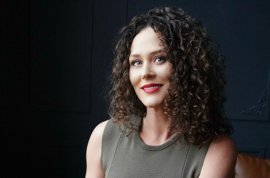 Woman with curly hair and red lipstick smiles in a dimly lit room. Wearing a sleeveless olive top. Dark, subtle background adds contrast.