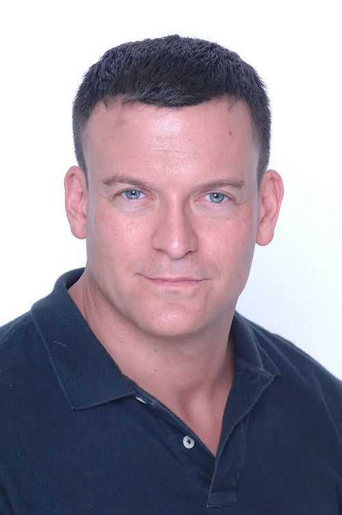 A person with short dark hair in a dark collared shirt looks directly at the camera against a plain white background.