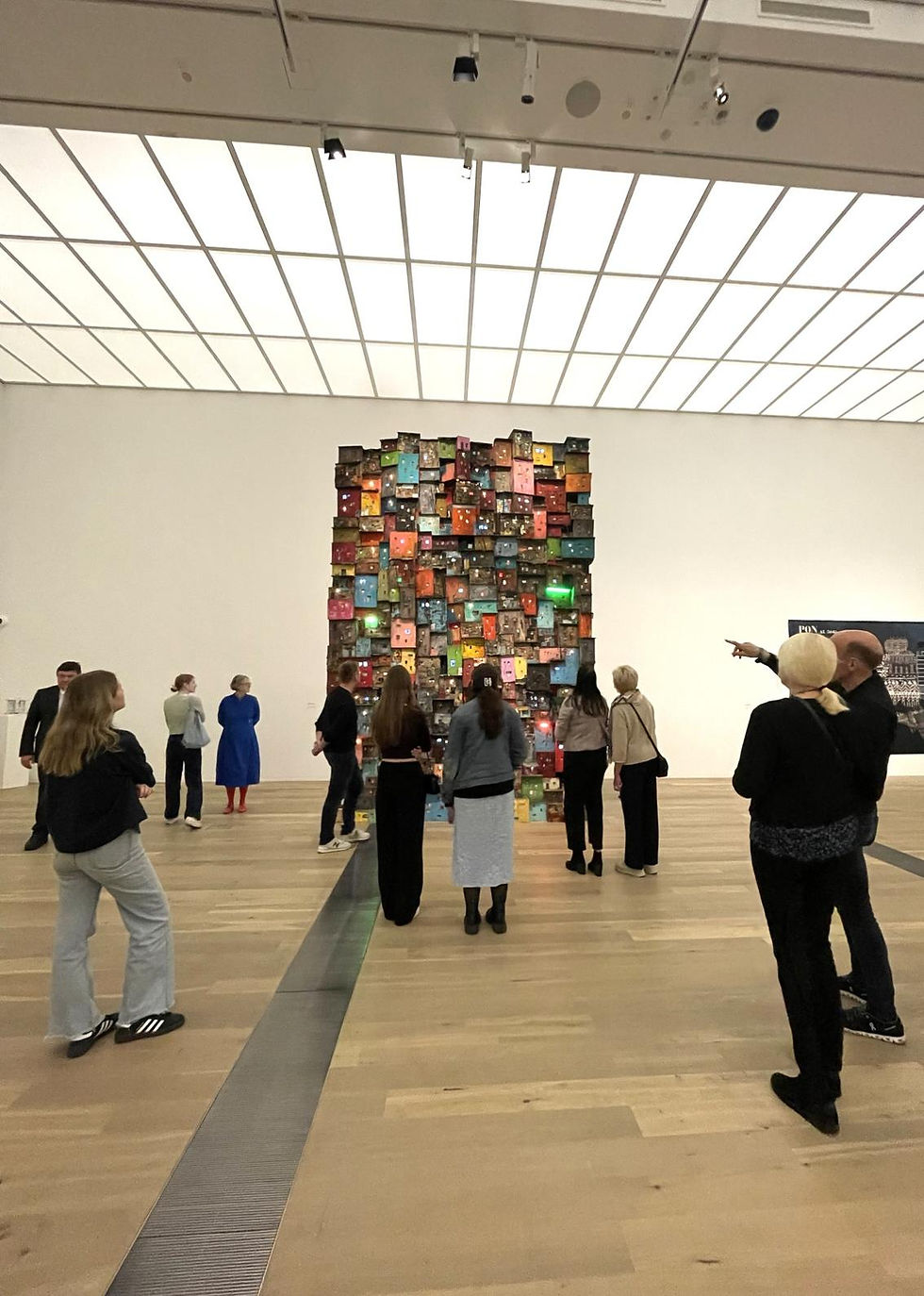 People in a gallery admire a tall, colorful installation of stacked boxes. Bright lights from above illuminate the scene.