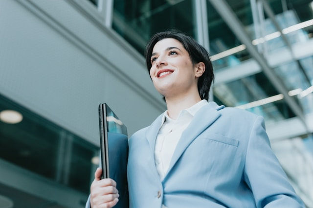 A confident woman in a light blue suit smiles while holding a laptop, walking through a modern glass office building.
