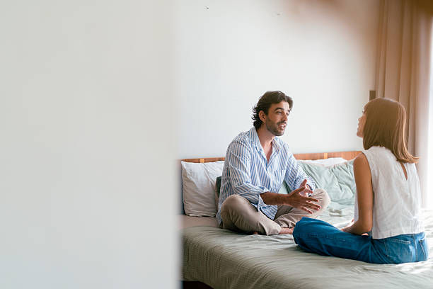 A man and woman sit facing each other on a bed in a bright bedroom, talking calmly and attentively.