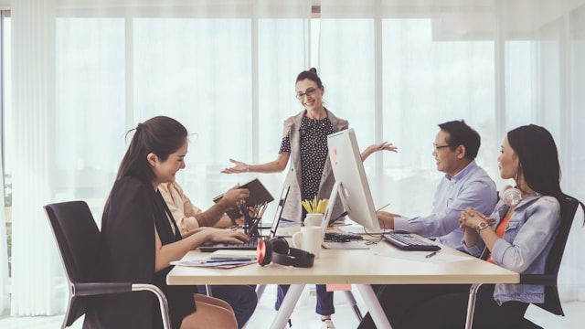 Five colleagues in a modern office engage in a lively discussion around a table. Computers and documents are present. Bright, airy setting.