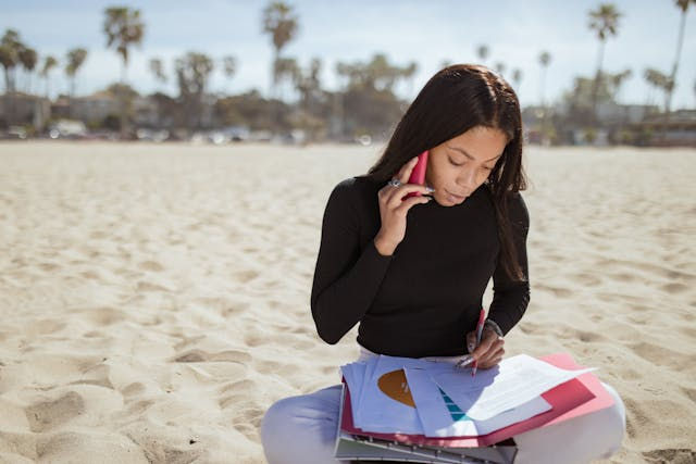 Woman sitting on a sandy beach, talking on a pink phone while reviewing documents with charts. Palm trees in the background, calm mood.