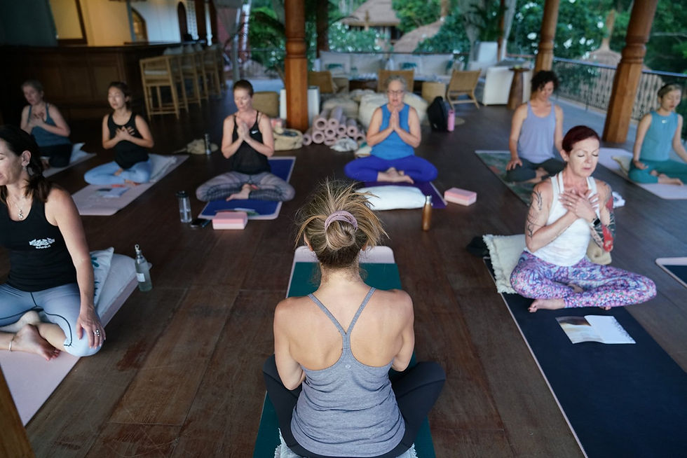 People in a yoga class meditate on mats with hands clasped, on a wooden floor. Bright colors, serene atmosphere, lush greenery outside.