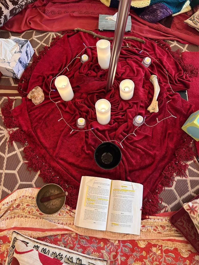 Ritual circle on a red blanket with lit candles, stones, a singing bowl and open book on patterned rugs, creating a calm mood