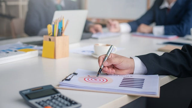 The photo shows a business meeting with individuals discussing documents, with one person marking a target symbol on a report.