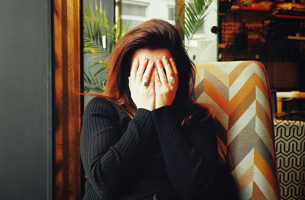 Woman in a black sweater covers her face with both hands in a patterned chair by a window, looking shy or upset.