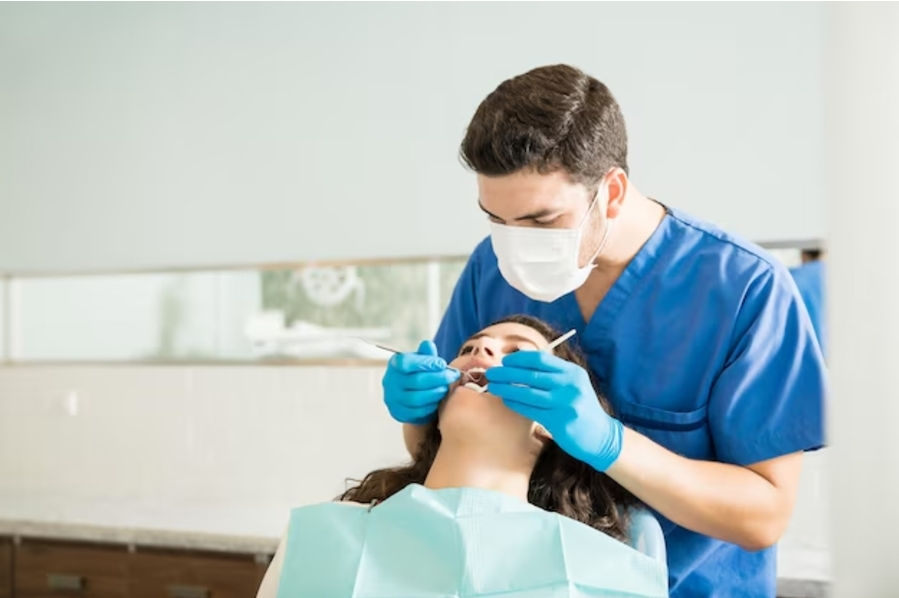 Dentist in blue scrubs and mask examines patient's teeth with tools in a clinic. Patient is reclined, wearing a dental bib.