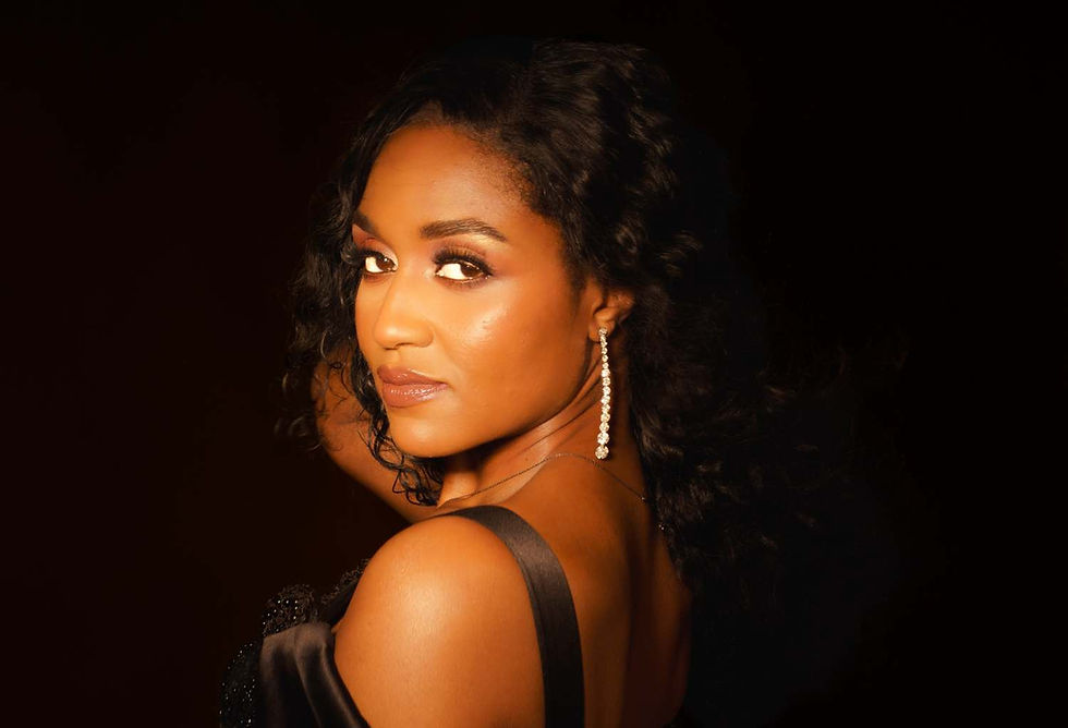 Woman with curly hair and long earrings poses in a dark setting. She wears a black off-shoulder dress and gazes over her shoulder.