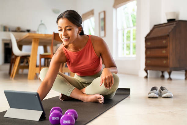 Woman in a red top sits on a yoga mat in a bright room, smiling while using a tablet. Purple dumbbells and shoes are nearby.