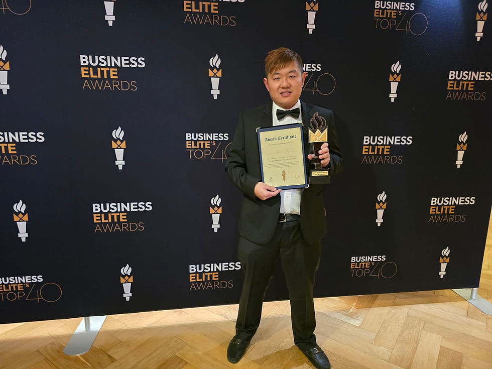 Man in a tuxedo holds an award certificate and trophy. He's standing in front of a "Business Elite Awards" backdrop with a wood floor.