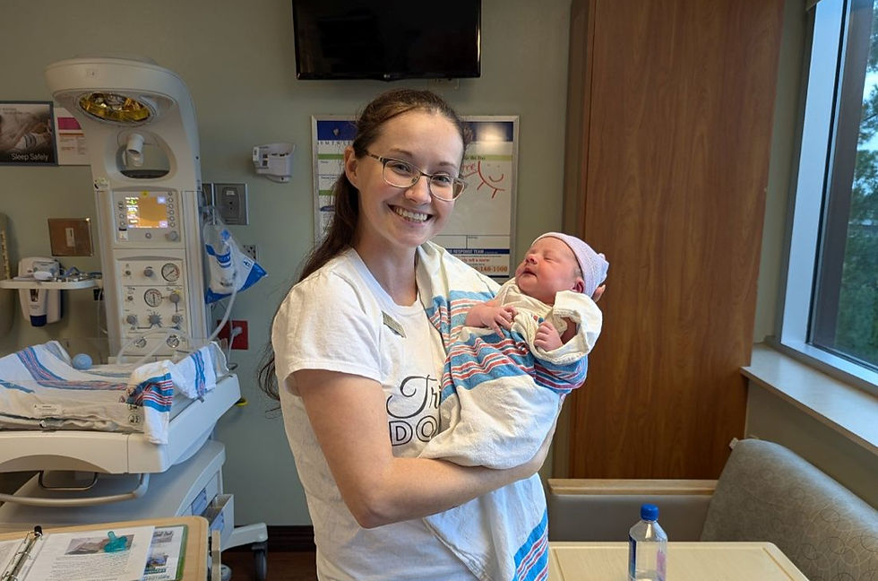 Smiling woman in a hospital room cradles a newborn wrapped in a striped blanket beside neonatal equipment.