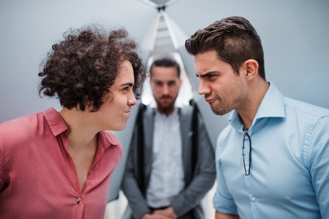 Two men stare angrily at each other in an office, with a blurred man standing behind them, creating a tense mood.